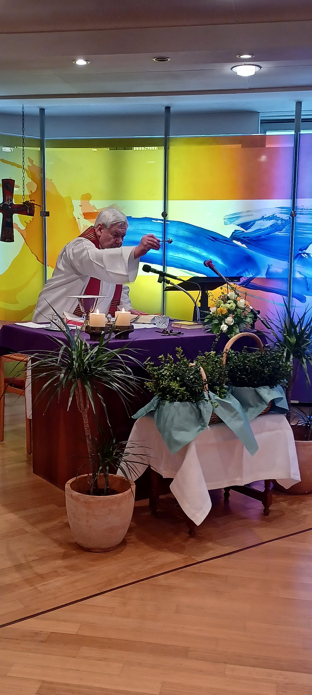 Person in weißer Kleidung steht an einem Tisch mit Mikrofonen und Blumenarrangements vor einer farbigen Glaswand.