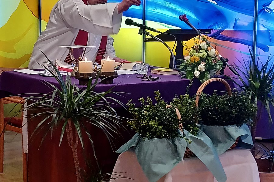 Person in weißer Kleidung steht an einem Tisch mit Mikrofonen und Blumenarrangements vor einer farbigen Glaswand.