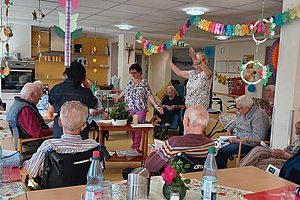 Mehrere ältere Menschen sitzen in einem Raum mit Tischen und Wasserflaschen, während einige Personen in der Mitte tanzen oder sich bewegen, der Raum ist mit bunten Girlanden dekoriert