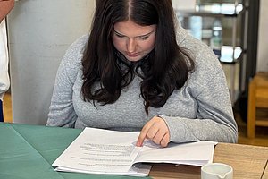 Person mit langen dunklen Haaren sitzt an einem Holztisch und liest in einem aufgeschlagenen Buch neben einer weißen Tasse