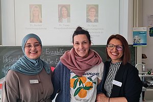 Drei Frauen stehen lächelnd zusammen, eine trägt ein Kopftuch, im Hintergrund ein Präsentationsschirm.