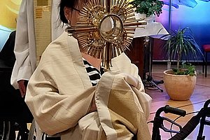 Person in Rollstuhl hält ein goldfarbenes Monstranzobjekt vor dem Gesicht, im Hintergrund steht eine weitere Person in liturgischer Kleidung vor einem bunt beleuchteten Fenster mit Kreuz und Pflanzen