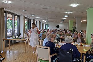 Mehrere ältere Menschen sitzen an Tischen in einem hellen Gemeinschaftsraum, eine Frau in weißem Kleid steht in der Mitte des Raums