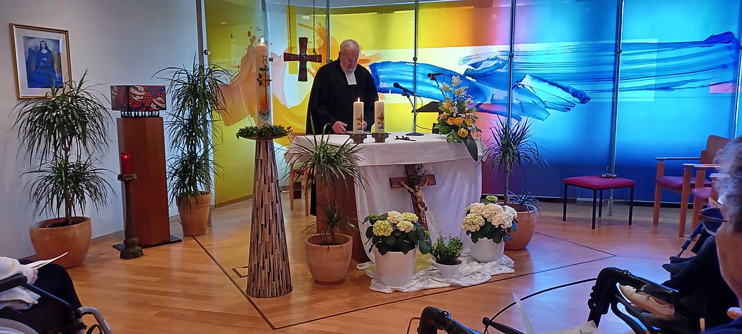 Ein Priester steht an einem mit Kerzen und Blumen geschmückten Altar in einer Kirche mit farbigem Glasfenster im Hintergrund, während mehrere Personen auf Stühlen sitzen.