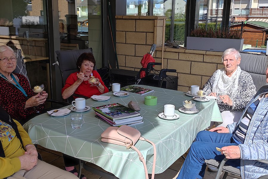 Sechs ältere Personen sitzen an einem rechteckigen Gartentisch mit Tassen, Tellern und Kuchen in einem überdachten Außenbereich.