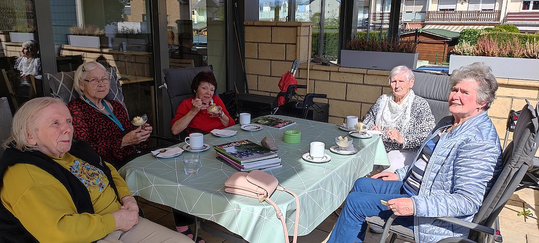 Sechs ältere Personen sitzen an einem rechteckigen Gartentisch mit Tassen, Tellern und Kuchen in einem überdachten Außenbereich.
