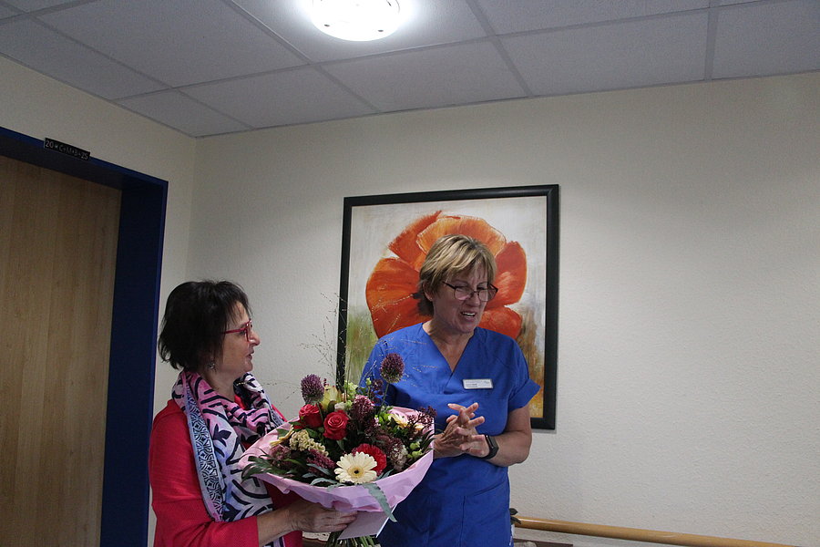 Frau in blauer Arbeitskleidung steht vor einem Bild mit roter Blume an der Wand, neben ihr Frau mit buntem Schal hält einen Blumenstrauß.