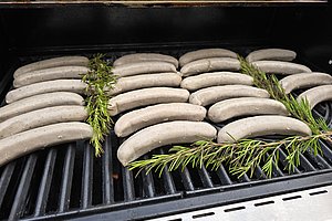 Mehrere helle Bratwürste liegen nebeneinander auf einem Grillrost mit Rosmarinzweigen dazwischen.