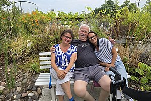 Drei Personen sitzen eng nebeneinander auf einer weißen Bank in einem Garten, die mittlere Person umarmt die beiden anderen, rechts neben ihnen steht ein Rollator