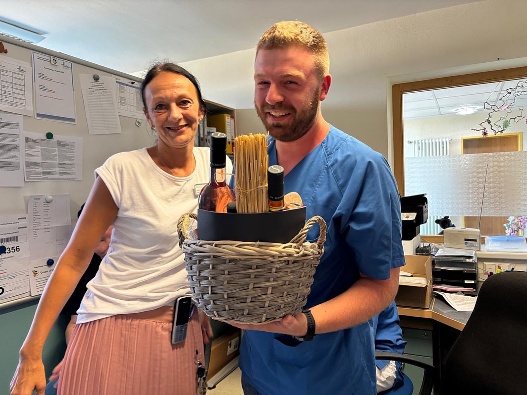 Zwei Personen in einem Büro, eine hält einen Korb mit Weinflaschen und Spaghetti, im Hintergrund Pinnwand und Schreibtisch mit Büroausstattung