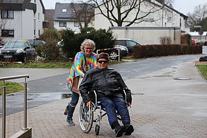 Frau schiebt einen Mann im Rollstuhl auf einem Gehweg, umgeben von Wohnhäusern und Bäumen.