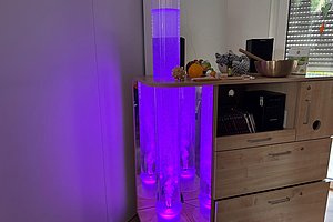 Leuchtende, zylindrische Glasgefäße mit violettem Licht auf einem Holzsideboard in einem Raum mit Fenster und geschlossener Jalousie