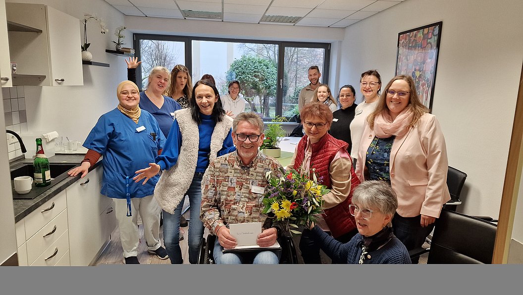 Gruppe von Menschen in einem hellen Raum mit Küchenzeile, eine Person im Rollstuhl hält ein Buch, eine andere überreicht einen Blumenstrauß.