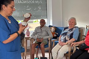 Pflegekraft in blauer Arbeitskleidung zeigt zwei runde Gegenstände vor einer Gruppe älterer Menschen, die auf Stühlen sitzen.
