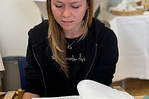 Frau mit langen Haaren sitzt an einem Tisch mit aufgeschlagenem Buch und mehreren Papieren