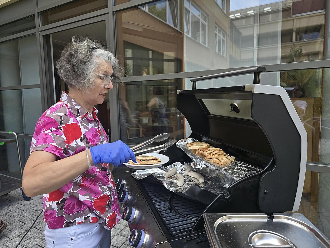 Ältere Frau mit grauen Haaren und blauem Handschuh wendet mit Zange Fischfilets auf einem Gasgrill auf einer Terrasse vor einem Gebäude