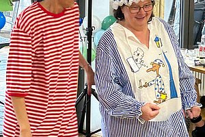 Zwei Frauen stehen nebeneinander, eine trägt ein rot-weiß gestreiftes Kleid, die andere eine blau-weiß gestreifte Schürze mit einem Entenmotiv und eine blaue Haube mit weißen Punkten