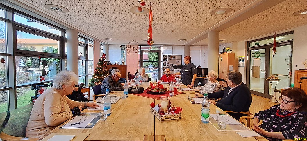 Mehrere ältere Menschen sitzen an einem langen Holztisch in einem hellen Raum mit Weihnachtsdekoration und sprechen miteinander.