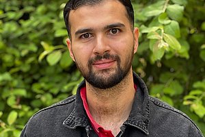 Mann mit dunklem Jeanshemd über rotem Hemd vor grünem Laub im Freien