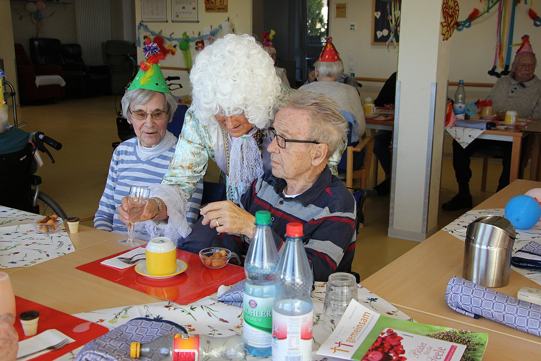 Person in weißer Perücke und Kostüm hält die Hände eines älteren Mannes mit grauen Haaren, beide sitzen an einem Tisch mit Getränken und Dekoration, im Hintergrund weitere Personen mit Partyhüten.