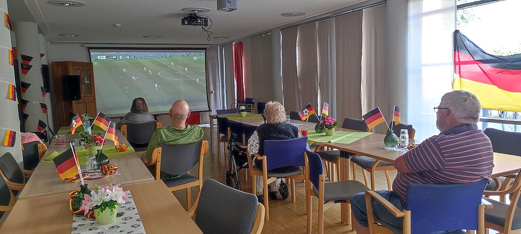 Mehrere ältere Menschen sitzen in einem Raum mit Tischen, auf denen kleine deutsche Fahnen stehen, und schauen auf eine Leinwand mit einem Fußballspiel.