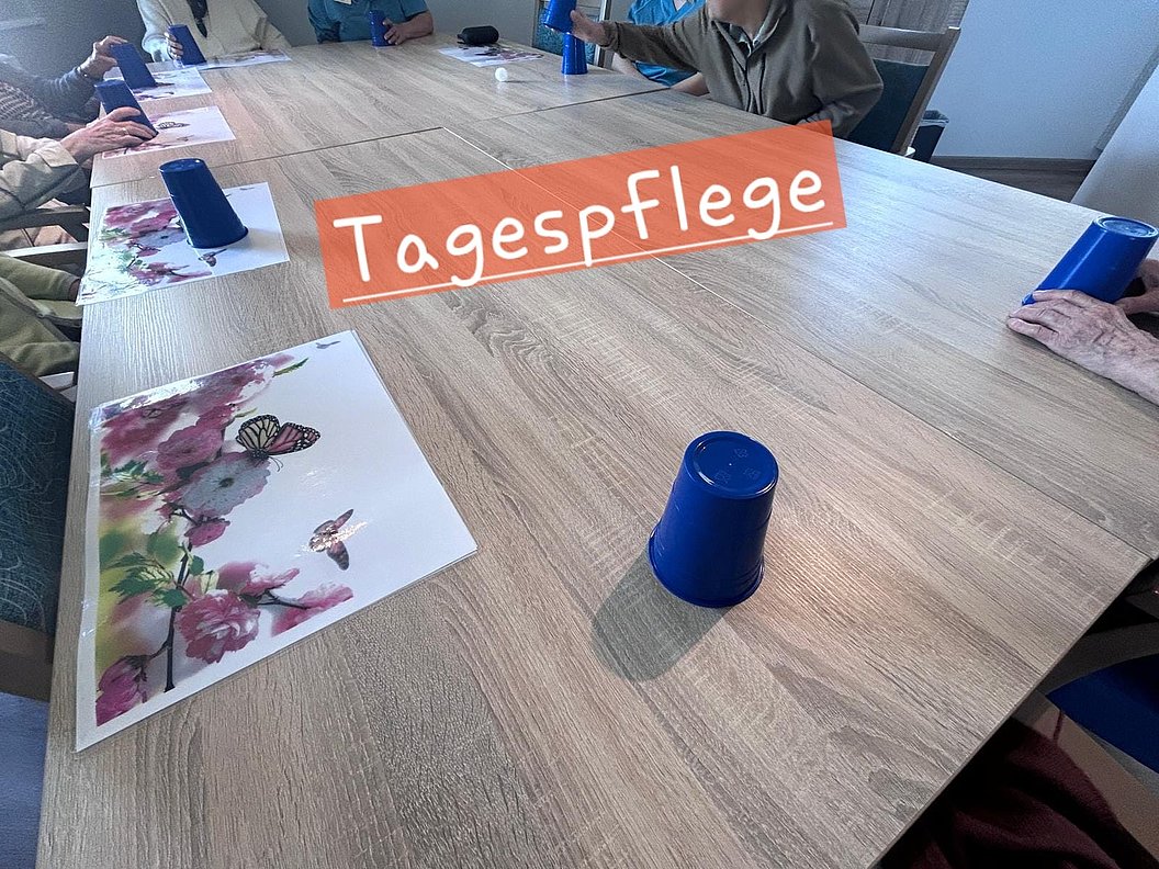 Mehrere ältere Menschen sitzen an einem Holztisch mit bunten Papieren und blauen Bechern darauf, darüber der Schriftzug 'Tagespflege'