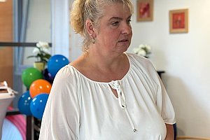 Frau mit hochgebundenem Haar und weißer Bluse steht in einem Raum mit bunten Luftballons und Bildern an der Wand