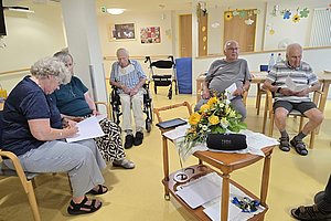 Ältere Menschen sitzen in einem Stuhlkreis in einem hellen Raum, einige lesen oder schreiben, ein Rollstuhl steht im Hintergrund