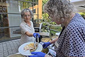 Zwei Frauen mit blauen Handschuhen servieren Essen auf einem Teller im Außenbereich eines Gebäudes