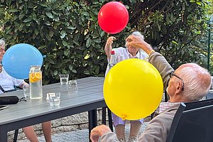 Mehrere ältere Menschen sitzen an einem Gartentisch und halten bunte Luftballons in den Händen