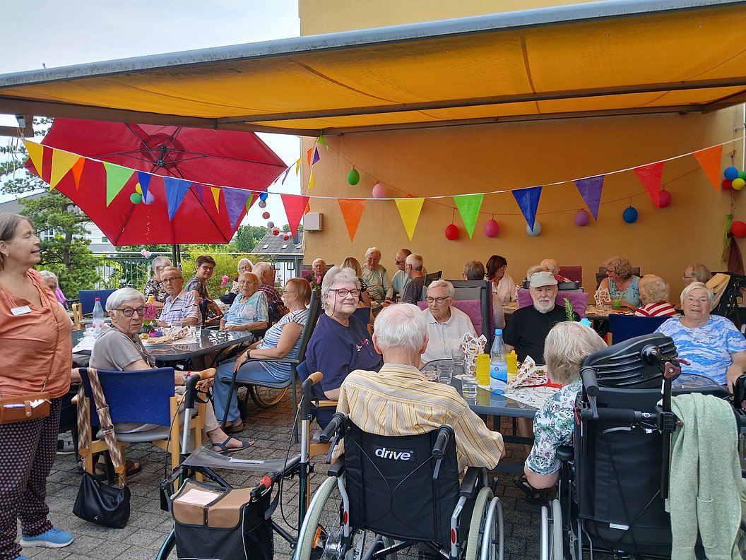 Gruppe älterer Menschen sitzt an mehreren Tischen unter einem gelben Sonnensegel mit bunten Wimpeln und Luftballons, einige im Rollstuhl, im Außenbereich eines Gebäudes