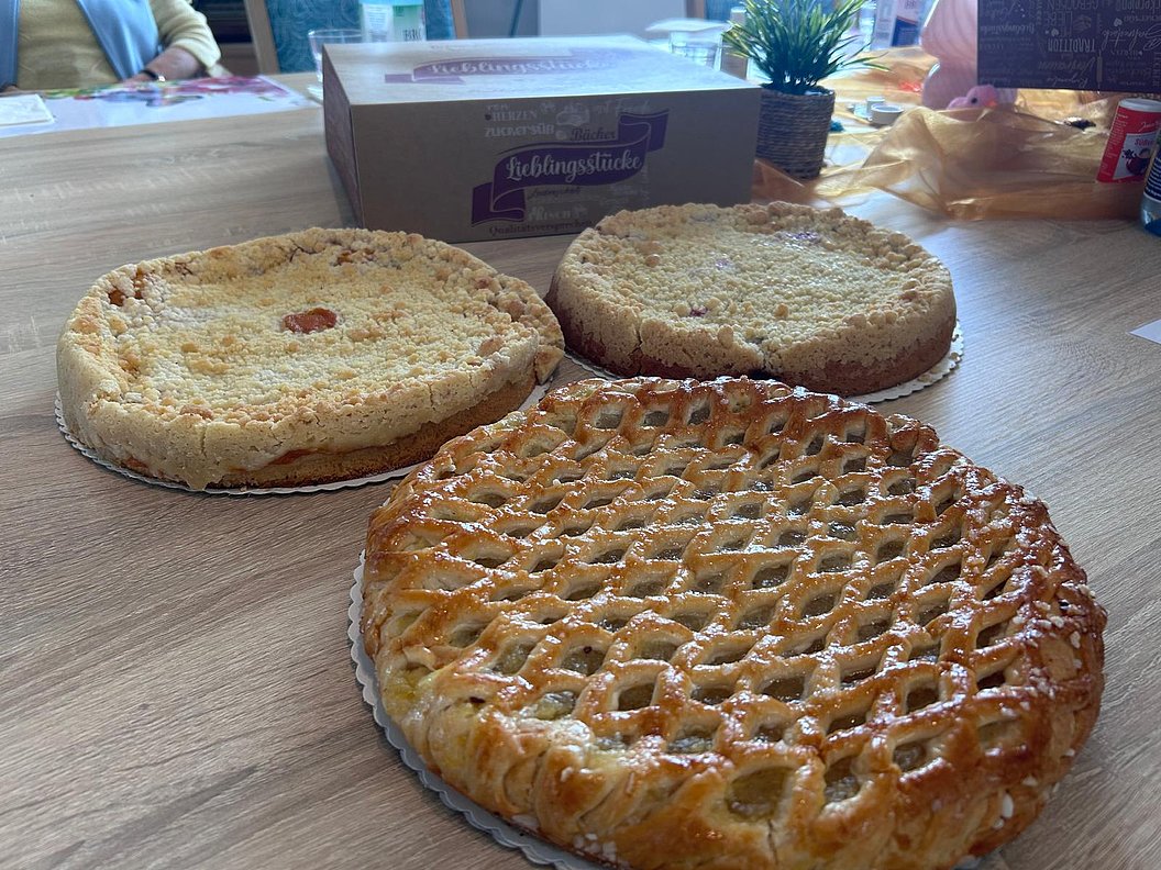 Drei verschiedene Kuchen auf einem Holztisch, im Hintergrund eine weiße Verpackung mit der Aufschrift 'Lieblingsstücke' und eine grüne Pflanze
