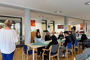 Frau steht vor Gruppe von Menschen, die an Tischen sitzen und ihr zuhören