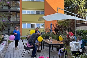 Mehrere Personen sitzen und stehen mit bunten Luftballons an einem Tisch vor einem mehrstöckigen Wohngebäude mit orange-gelben Fassadenabschnitten