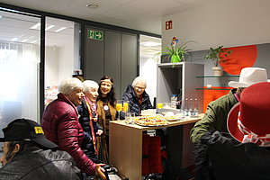 Gruppe von Menschen steht um einen Tisch mit Getränken und Snacks in einem modernen Büro mit grauen Schränken und orangefarbenen Wandelementen