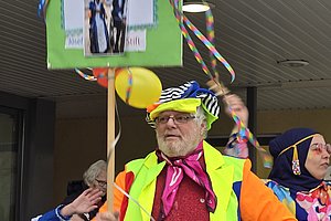Mann in buntem Kostüm mit Luftballons und Schild bei einer Festlichkeit, lebendige Karnevalsstimmung.