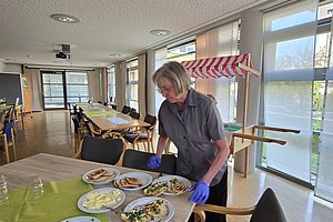 Frau mit blauen Handschuhen steht an einem Tisch mit mehreren Tellern mit verschiedenen Speisen in einem hellen Raum mit großen Fenstern
