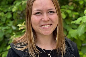 Junge Frau mit langen blonden Haaren trägt schwarzen Kapuzenpullover und schwarzes T-Shirt mit weißer Schrift vor grünem Laub im Hintergrund