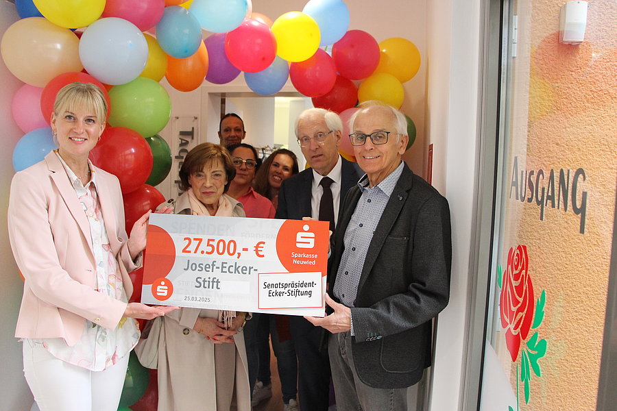 Mehrere Personen stehen unter einem Bogen aus bunten Luftballons und halten gemeinsam einen großen symbolischen Scheck mit der Aufschrift '27.500,- € Josef-Ecker-Stift'.