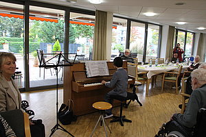 Person spielt Klavier in einem Raum mit großen Fenstern, mehrere ältere Menschen sitzen an Tischen und hören zu