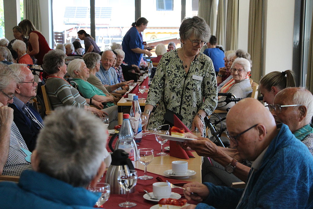 Senioren sitzen an einem langen Tisch in einem Gemeinschaftsraum, eine Frau serviert Essen