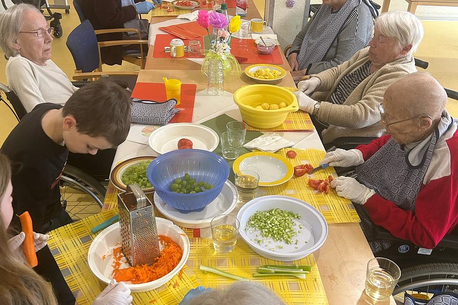 Mehrere ältere Menschen sitzen an einem Tisch und schneiden Gemüse, während eine jüngere Person mit Handschuhen eine Reibe benutzt.