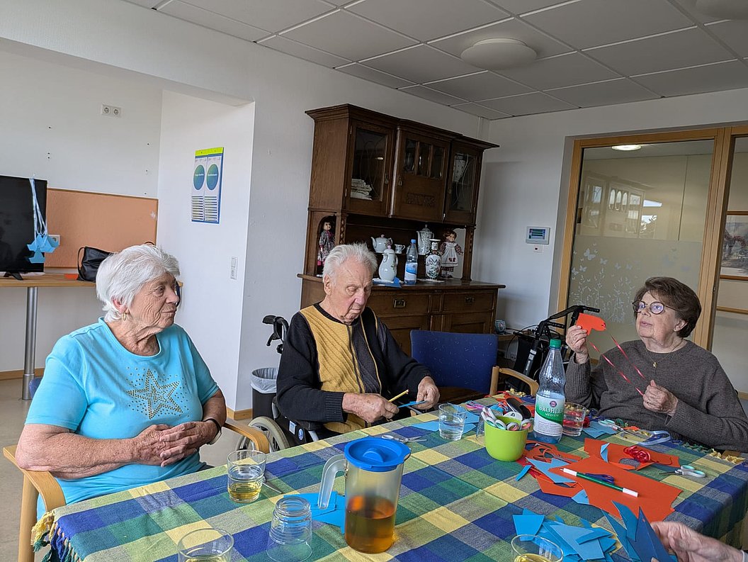 Drei ältere Personen sitzen an einem Tisch mit kariertem Tischtuch, auf dem Bastelmaterialien und Getränke stehen.