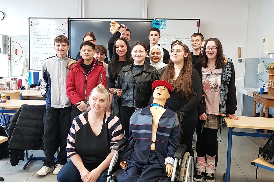 Gruppe von Menschen in einem Klassenraum, eine Person sitzt im Rollstuhl mit einer Puppe bekleidet in einem blauen Anzug und roter Mütze.