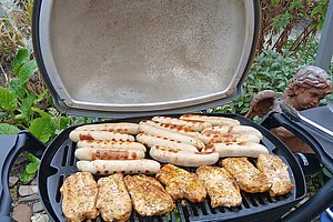 Grill mit offenem Deckel und mehreren Würstchen und marinierten Fleischstücken auf dem Grillrost im Garten