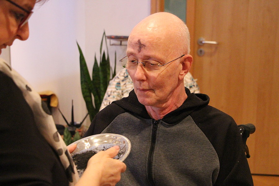 Ein älterer Mann mit grauem Kapuzenpullover sitzt vor einer geschlossenen Holztür, eine Person reicht ihm einen Teller mit Essen.
