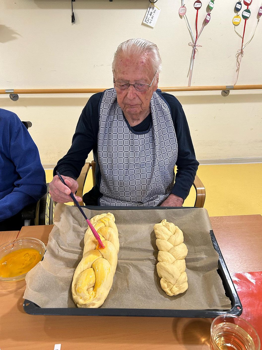 Person mit Schürze bestreicht zwei geflochtene Teigstücke auf einem Backblech mit Backpapier.