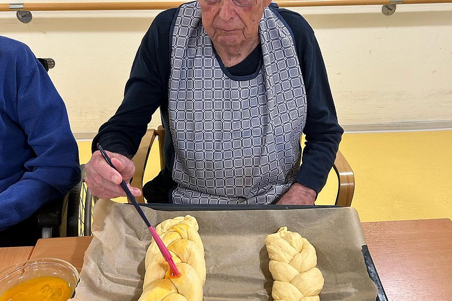 Person mit Schürze bestreicht zwei geflochtene Teigstücke auf einem Backblech mit Backpapier.