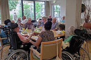 Gruppe älterer Menschen sitzt an mehreren Tischen in einem hellen Gemeinschaftsraum mit großen Fenstern