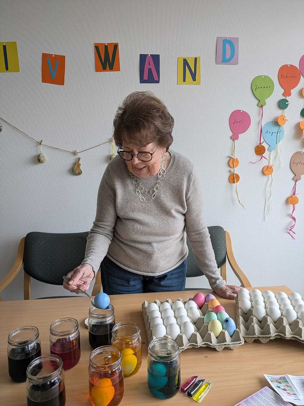 Person steht an einem Tisch mit mehreren Gläsern gefärbter Flüssigkeit, Eierkartons mit gefärbten Eiern und bunten Buchstaben an der Wand.